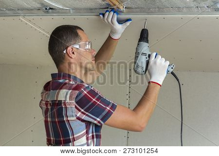 Young Man In Goggles Fixing Drywall Suspended Ceiling To Metal Frame Using Electrical Screwdriver On