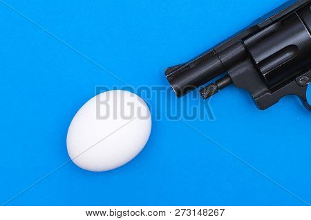 White Chicken Egg And Black Handgun Weapon Isolated On Blue Background.