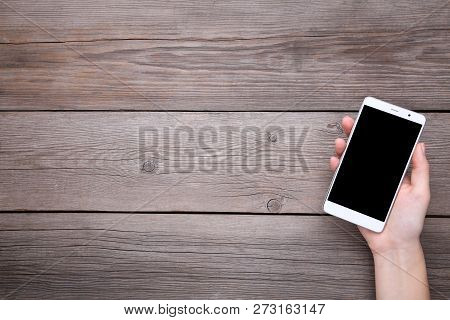 Female Hands Holding Mobile Phone With Blank Screen On Grey Wooden Table. Smartfone White
