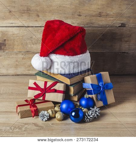 A Stack Of Books, Santa Clauss Cap, Cones, Christmas-tree Decorations, Gift Boxes On A Wooden Backgr