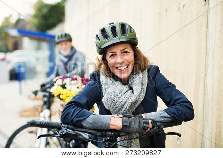 Active Senior Couple With Electrobikes Standing Outdoors On A Pathway In Town.