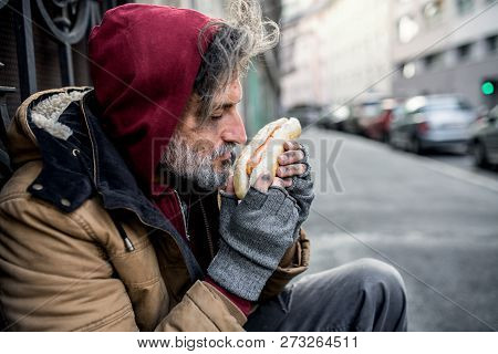 Homeless Beggar Man Outdoors In City, Holding And Smelling Hot-dog.