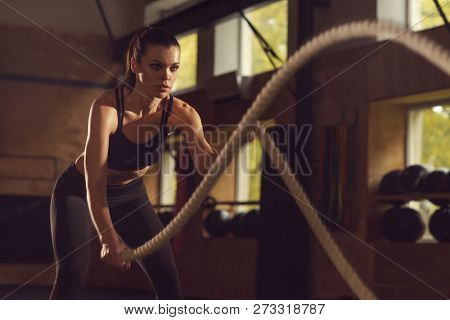 Fit, sporty and athletic sportswoman working in a gym. Woman training using battle ropes. Sports, athletics and fitness concept.