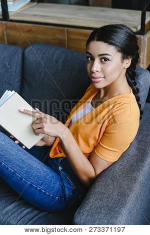 High Angle View Of Beautiful Mixed Race Girl In Orange Shirt Holding Book And Looking At Camera In L