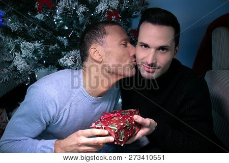 Gay Male Couple Exchanging Christmas Gift In Front Of Tree. One Kissing, Other Smiling At Camera