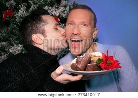 Male Gay Couple Kissing And Laughing In Front Of Christmas Tree With Plate Of Cakes