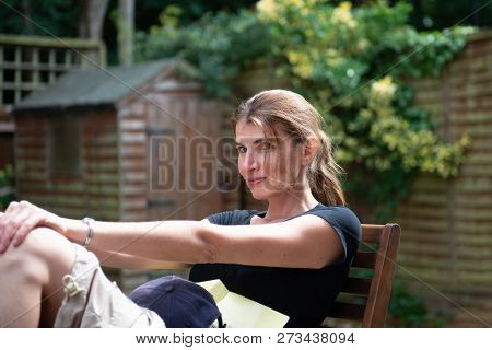Young Female Sitting In The Chair In The Garden With The Book And The Hat Looking At The Camera Smil