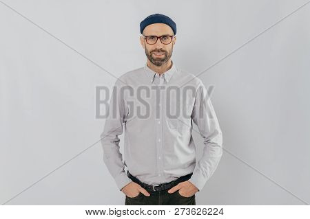 Indoor Shot Of Satisfied Caucasian Man In Spectacles With Bristle, Keeps Hands In Pockets, Dressed I