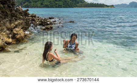 Mother And Daughter With Mobile Phone Sitting In Sea At Beach Surrounded By Tropical Fish