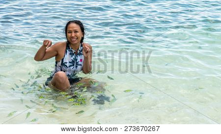 Asian Woman Sitting In Water At Beach, Laughging And Playing While Surrounded By Tropical Fish