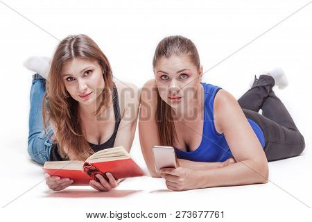 Two Young Attractive Caucasian Women Lye On The Floor Legs Up. One Holds A Book With Red Cover, The 