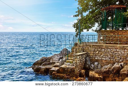 Lovran, Croatia. Rocky stone coastline with big rocks pavilion in shade of tree and stairs to Adriatic Sea. Blue waves sky clouds above horizon.