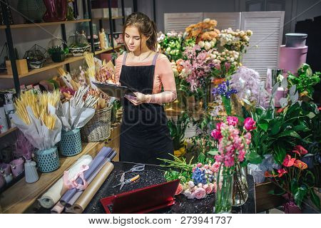 Busy Young Florist Stand At Table And Hold Black Planchette And Writedown. She Is Surround With Diff