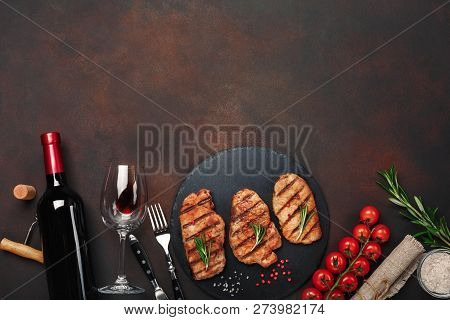 Grilled Pork Steaks On Stone With Bottle Of Wine, Wine Glass, Knife And Fork On Rusty Background. To