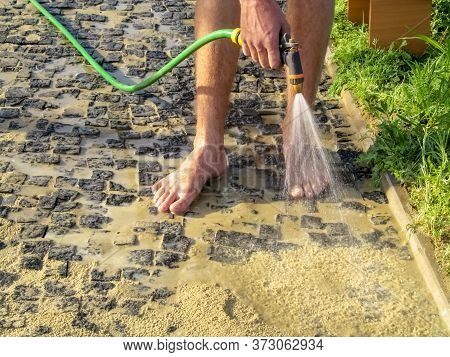 A Barefoot Worker Washes Sand From A Newly Laid Granite Stones With A Garden Hose With A Rain. Stage