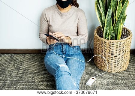 Young Woman Sitting Against A Wall Holding A Smartphone While Charging A Smartphone Battery.