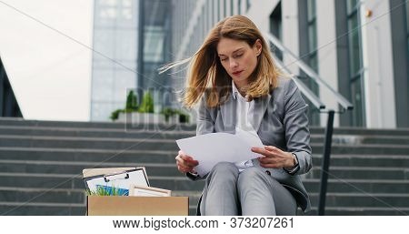 Fired Caucasian Femae Office Worker Sitting On Stairs In Depression With Box Of Stuff. Unemployed Bu
