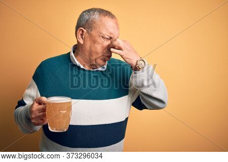 Senior handsome man drinking jar of beer standing over isolated yellow background smelling something stinky and disgusting, intolerable smell, holding breath with fingers on nose. Bad smell