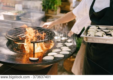 The Cook Roasts The Courgettes. Grilled Vegetables. Round Bowl-shaped Grill With A Fire Inside.