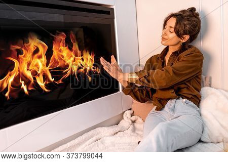 Young Woman Relaxing Near The Fireplace, Warming Hands At The Modern Living Room At Home