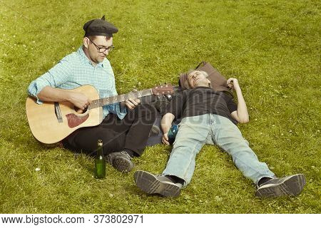 Two Men On City Park Summer Meadow Enjoying Day With Guitar
