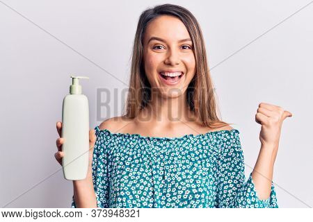 Young beautiful girl holding bottle of cream lotion pointing thumb up to the side smiling happy with open mouth 