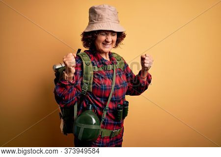 Middle age curly hair hiker woman hiking wearing backpack and water canteen using binoculars very happy and excited doing winner gesture with arms raised, smiling and screaming for success. Celebratio