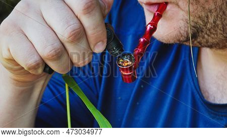 A Man Ignites Marijuana In A Pipe. A Guy In A Blue T-shirt Smokes Marijuana From A Beautiful Red Pip