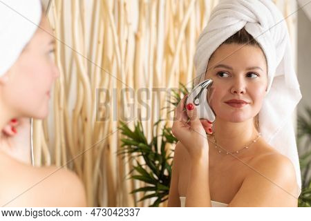 An Attractive Woman Does A Facial Massage With A Microcurrent Massager In Front Of A Bathroom Mirror