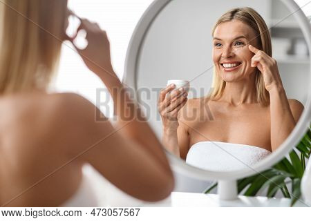 Anti-age Skincare. Beautiful Middle Aged Woman Applying Moisturising Eye Cream Near Mirror