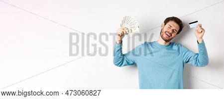 Handsome Man Dancing With Money And Plastic Credit Card, Standing In Casual Clothes Over White Backg