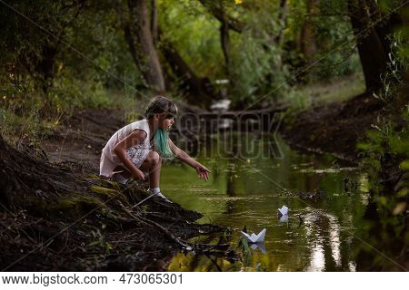 Teenage Girl With Green Hair In Dress, Stands On The River Bank, Launches White Paper Origami Boat