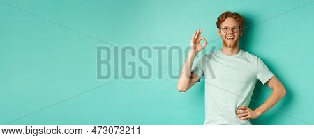 Happy Young Man With Red Hair And Beard, Wearing Glasses And T-shirt, Smiling Satisfied And Showing 