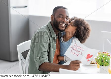 Curly African American Girl Kissing Cheek Of Happy Father With Greeting Card On Fathers Day.