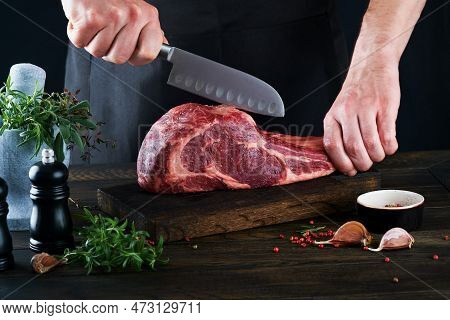 Chef Cutting Steak Beef. Mans Hands Hold Raw Steak Tomahawk On Rustic Wooden Cutting Board On Black 