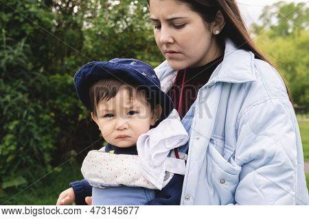 A Male Infant Is Looking At Camera While Being Carried By His Mother With A Baby Carrier, Wearing Bl