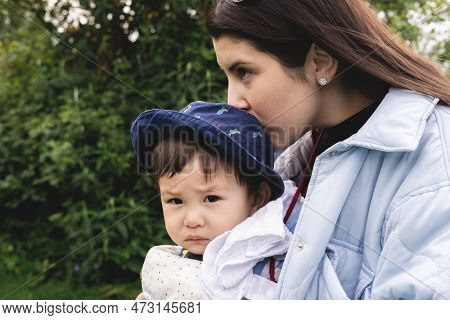A Male Infant Looking At Camera Being Carried With A Baby Carrier And Kissed On The Head By His Moth