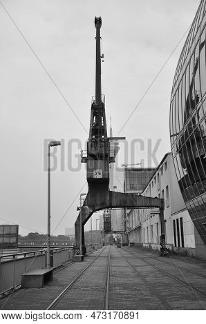 Vertical Grayscale Shot Of An Old Cargo Crane In The Media Harbor In Dusseldorf, Germany.