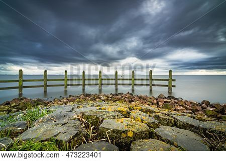 Lake View At Dark Time With Heavy Clouds And Stormy Winds. Stretch Dam In The Lake With A Wooden Pil