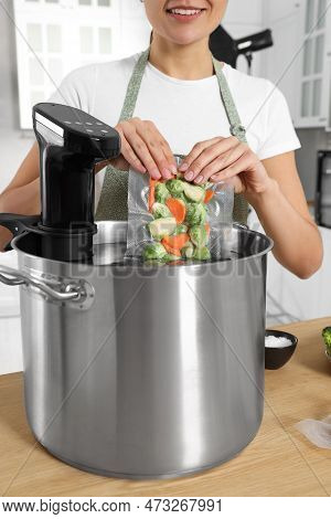Woman Putting Vacuum Packed Vegetables Into Pot With Sous Vide Cooker In Kitchen, Closeup. Thermal I