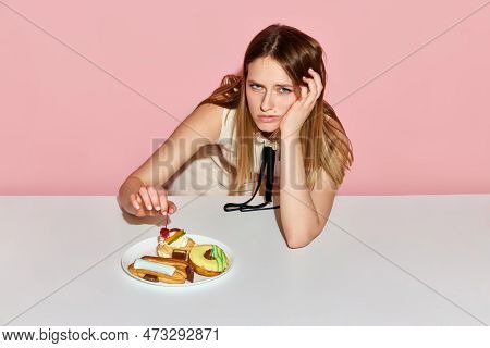 Upset Young Model Surrounded By Junk Food Looking At Camera With Sad Eyes Over Pink Background