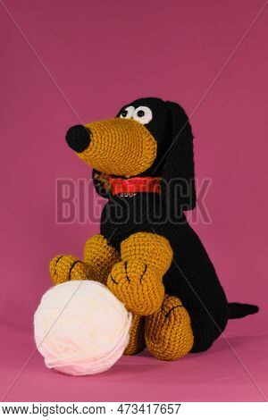 Amigurumi Dog Doll Play With Ball On Pink Background. A Soft Diy Toy Made Of Cotton. One Black And R