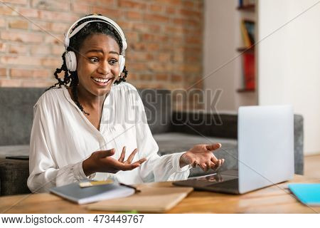 Video Call. Emotional Black Woman In Headphones Talking And Gesturing At Laptop Webcamera, Sitting A