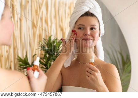 An Attractive Woman In Front Of A Mirror Applies A Serum To The Skin Of The Face In The Bathroom.
