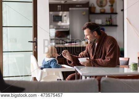 Father Wearing Bathrope Spoon Feeding Hir Infant Baby Boy Child Sitting In High Chair At The Dining 