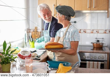 Cheerful Senior Woman Showing A Freshly Baked Homemade Plumcake At Her Husband Washing The Dishes. C
