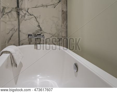White Acrylic Bathtub. Close-up. The Towel Is Thrown Over The Edge. On The Beige Marble Tile Wall Th