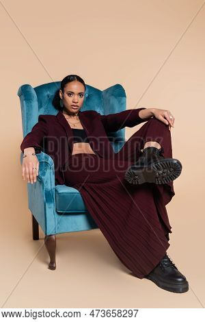Stylish African American Model In Maroon Blazer And Wide Pants Sitting On Blue Velvet Armchair On Be