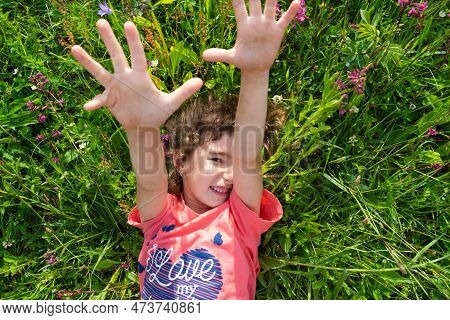 Portrait Of A Child Girl In Summer Lying In The Grass And Wildflowers With Heels And Palms. Summer T