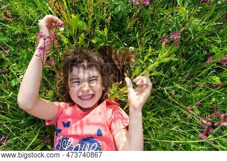 Portrait Of A Child Girl In Summer Lying In The Grass And Wildflowers With Heels And Palms. Summer T